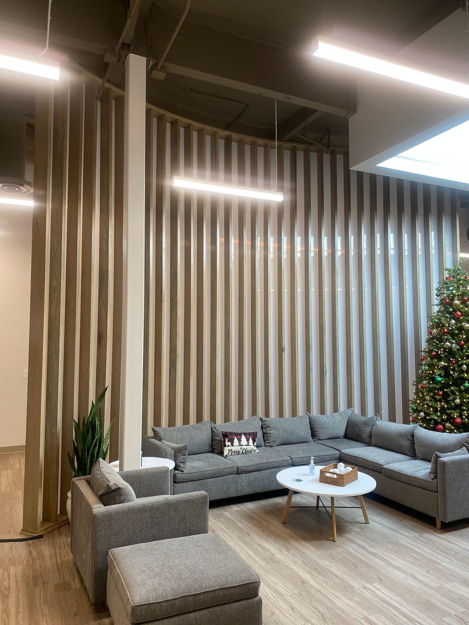 IMG_4243 Modern office lounge area with a gray sectional sofa, wooden slatted walls, and hanging linear lights. a christmas tree is visible in the background.